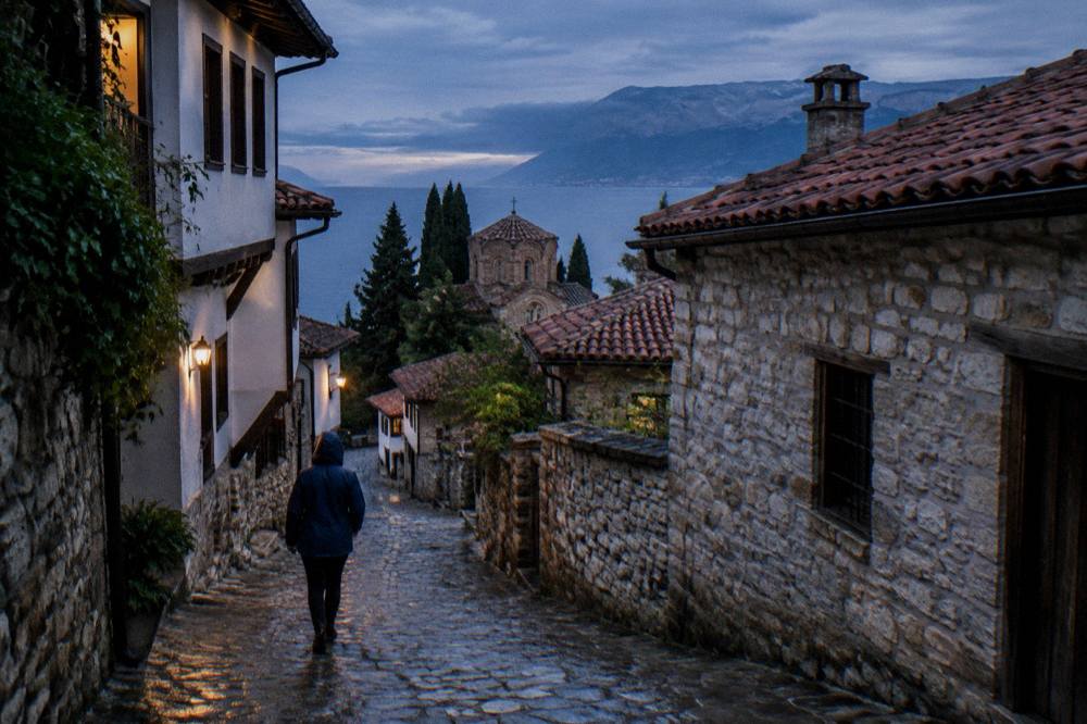 Person with a rain jacket walking a narrow wet street in Ohrid past stone houses