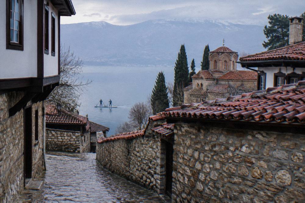 Two people on a tandem paddleboard passing the stone walls and red roofs of Ohrid old town