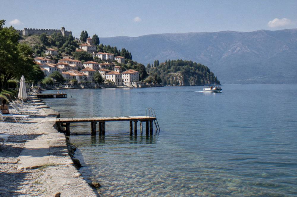 A small tour boat crossing open blue water toward a wooded peninsula on Lake Ohrid