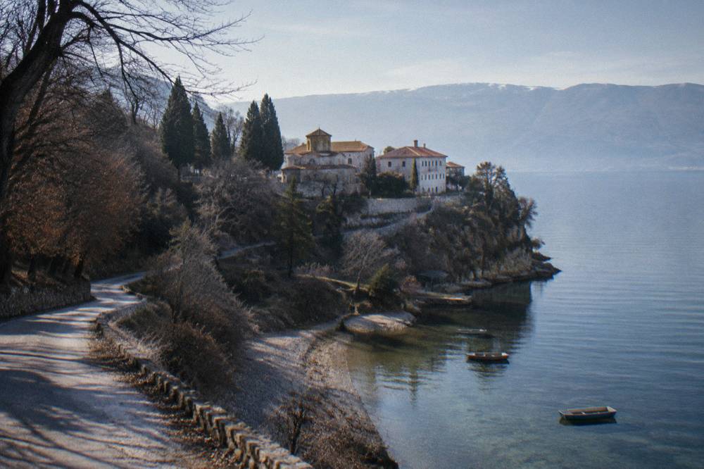 A narrow road winding along a wooded lakeshore with pebble beaches below